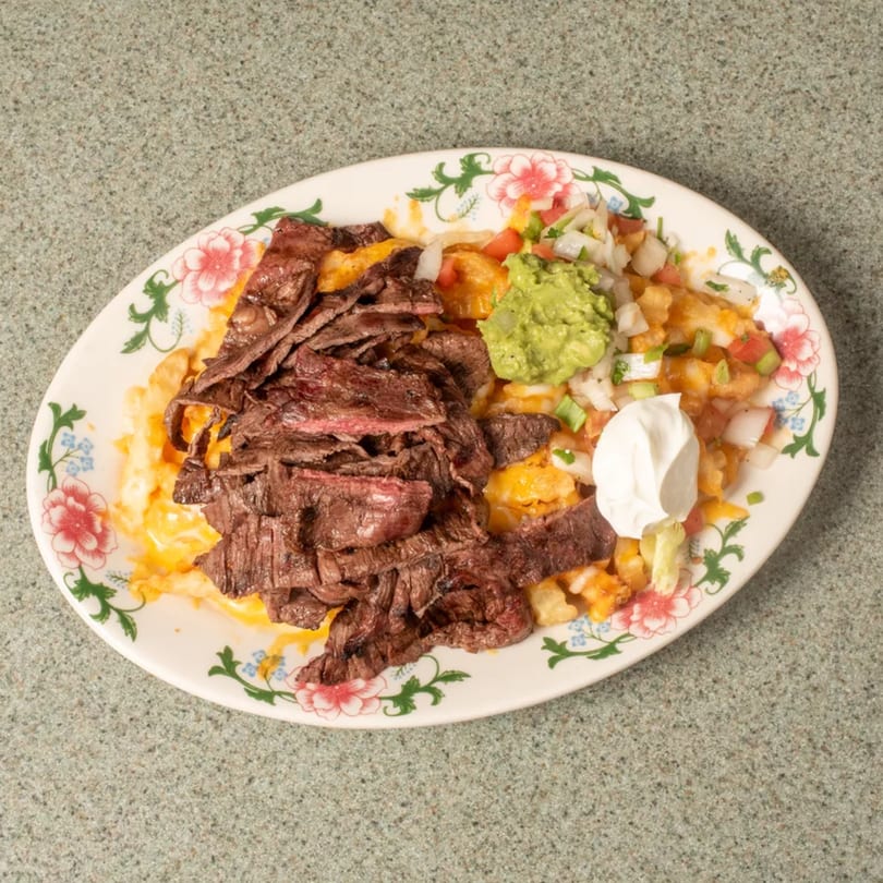 Sliced steak and cheesy fries with guacamole, pico de gallo, and sour cream on a floral plate.
