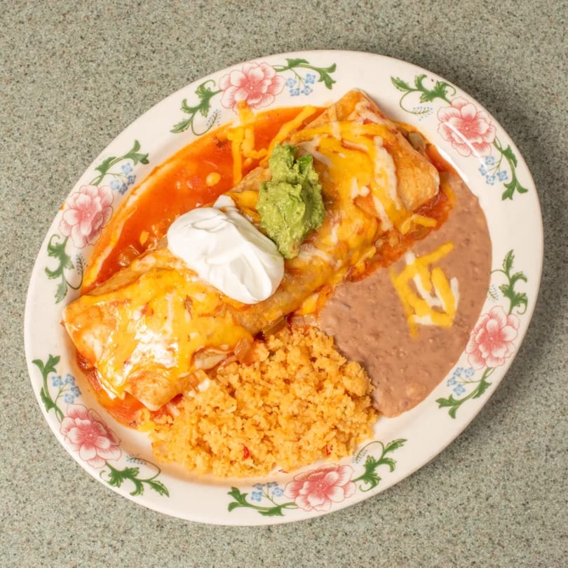 Two cheesy burritos with sour cream, guacamole, rice, and refried beans on a floral plate.