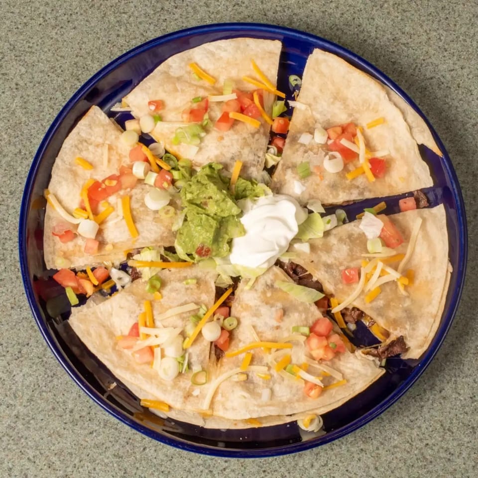 Quesadilla wedges on a blue plate, topped with guacamole, sour cream, cheese, tomatoes, and green onions.