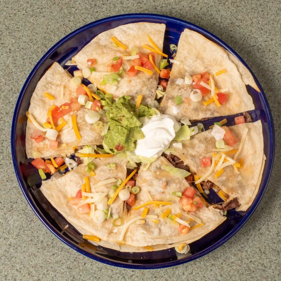 Quesadilla wedges topped with guacamole, sour cream, tomatoes, cheese, and green onions on a blue plate.