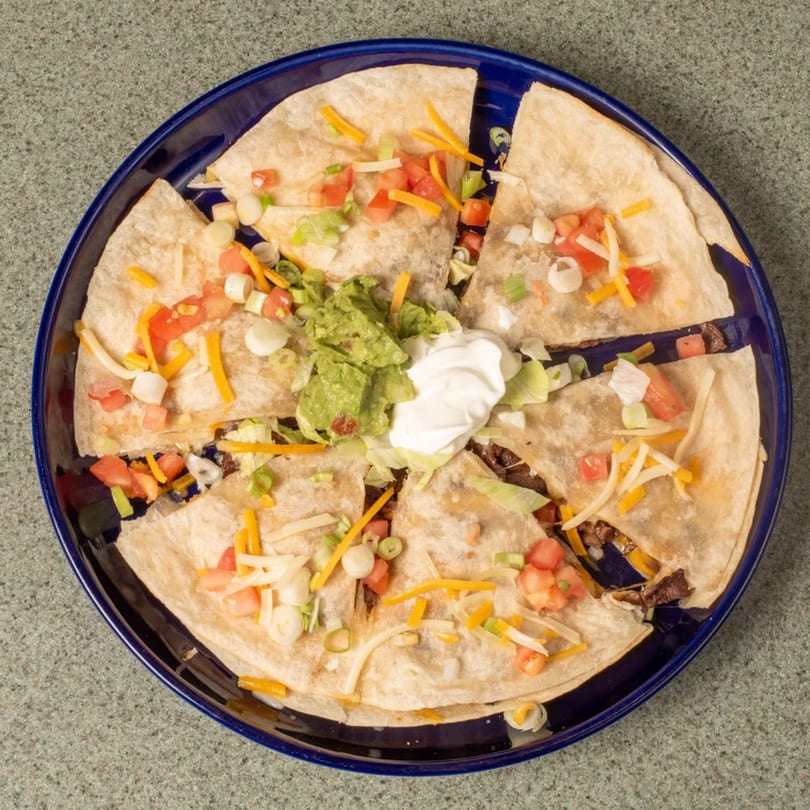 Quesadilla wedges topped with guacamole, sour cream, tomatoes, cheese, and green onions on a blue plate.
