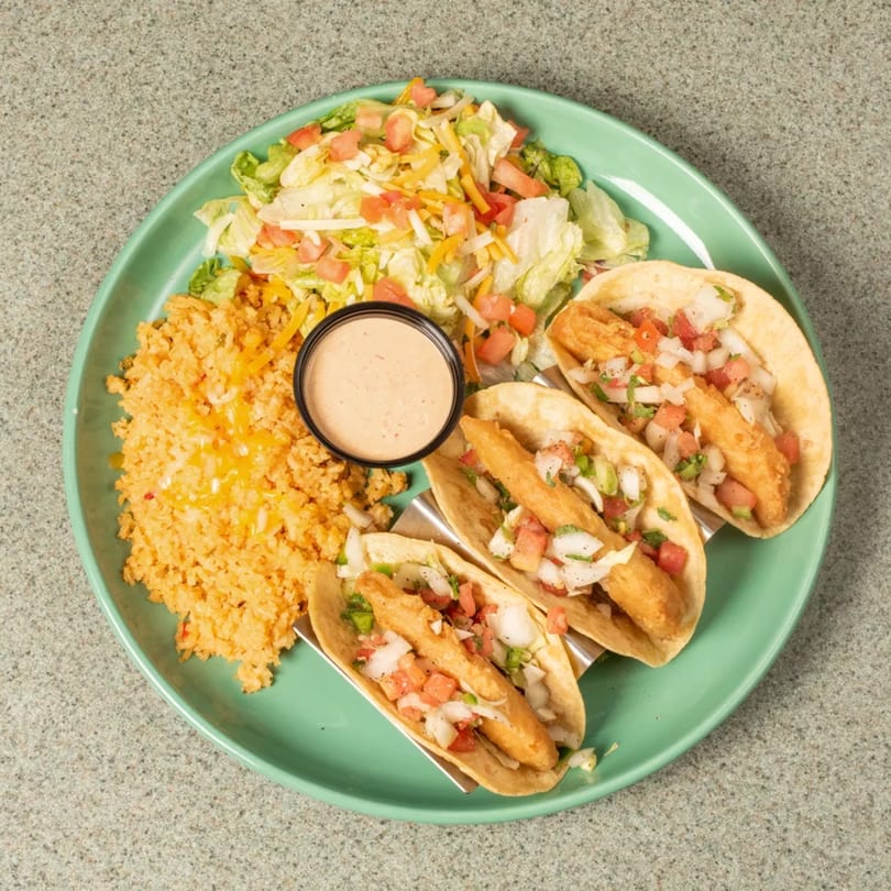 Three fish tacos with pico de gallo, served with rice, salad, and a side of sauce on a green plate.